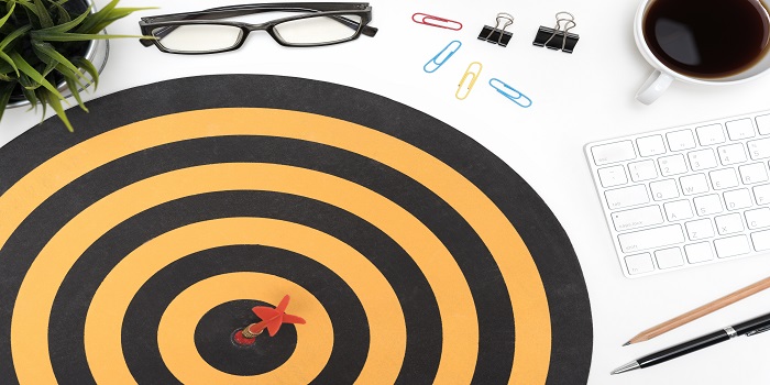 Dart target arrow hitting on bullseye in dartboard over office desk table background with eye glasses, pen, pencil, computer and cup of coffee